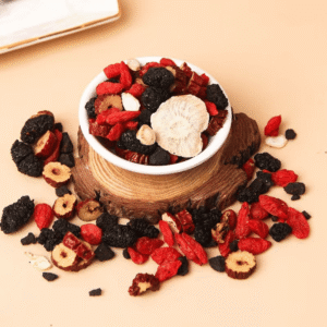 A white bowl filled with dried red dates, goji berries, mulberries, and ginseng slices resting on a wooden slice.